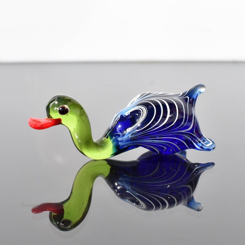 May include: A glass figurine shaped like a duck with a bright red beak, a translucent light green neck and head, and a deep blue body featuring swirling white glass patterns. The duck is positioned horizontally with a smooth, reflective finish.