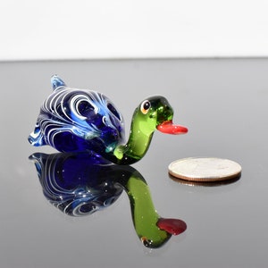 May include: A small glass duck figurine featuring a deep blue body with white swirling patterns, a translucent green neck, and a bright red beak. The duck is positioned next to a silver-colored coin for scale, showing its miniature size.
