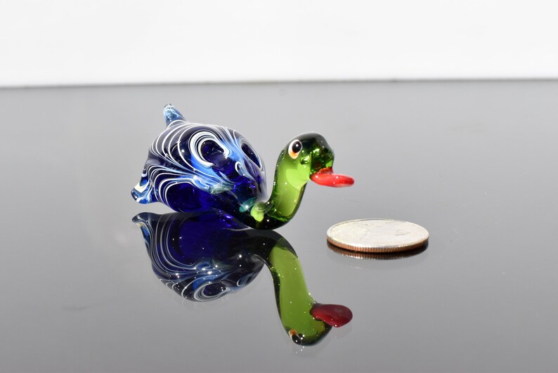 May include: A small glass duck figurine featuring a deep blue body with white swirling patterns, a translucent green neck, and a bright red beak. The duck is positioned next to a silver-colored coin for scale, showing its miniature size.