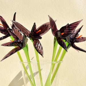 May include: A glass vase holds several dark purple glass flowers with orange centers and green stems. The flowers have pointed petals and are arranged in a clear glass vase, set against a light background.