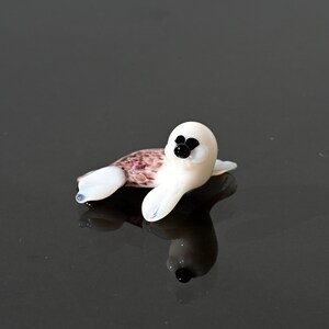 May include: A small, glass seal figurine. The seal has a white body with black eyes and a brown and white patterned back. The figurine is lying down on a reflective surface.