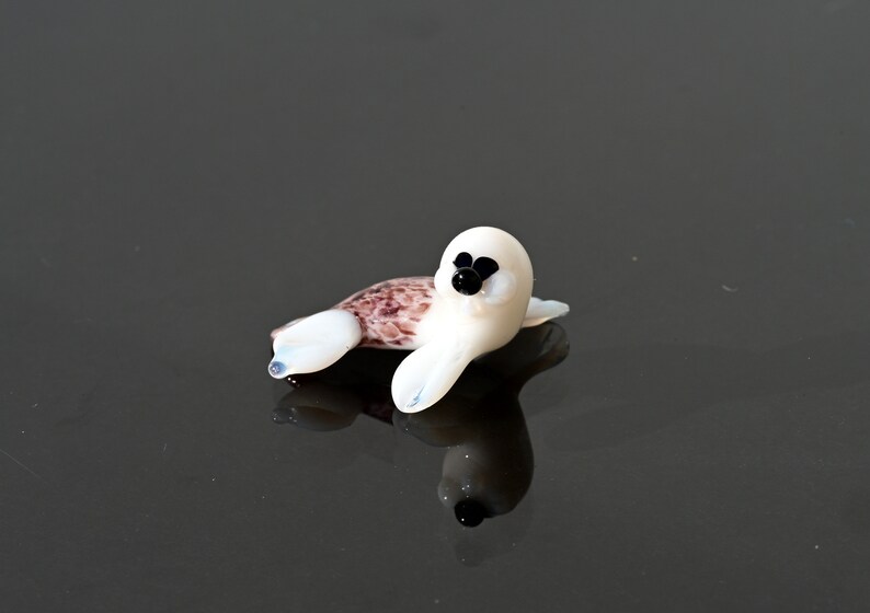 May include: A small, glass seal figurine. The seal has a white body with black eyes and a brown and white patterned back. The figurine is lying down on a reflective surface.