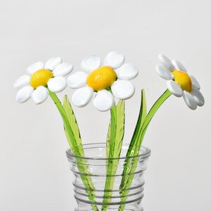 May include: Three white glass daisies with yellow centers in a clear glass vase. The daisies have green stems.