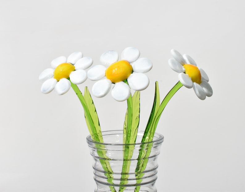 May include: Three white glass daisies with yellow centers in a clear glass vase. The daisies have green stems.