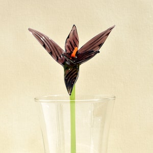 May include: A handcrafted glass flower with dark purple petals and an orange center, displayed in a clear glass vase. The flower has a green stem. The glass flower is a decorative item.
