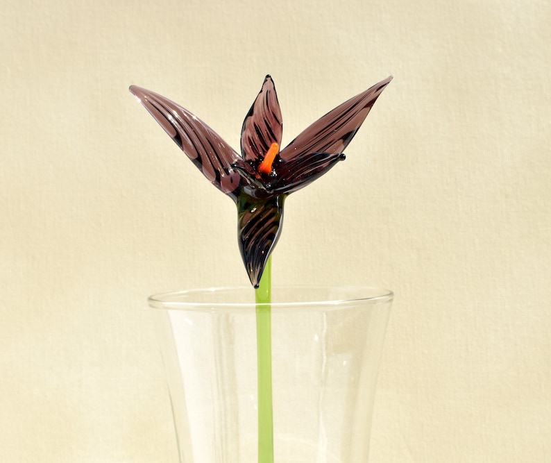 May include: A handcrafted glass flower with dark purple petals and an orange center, displayed in a clear glass vase. The flower has a green stem. The glass flower is a decorative item.