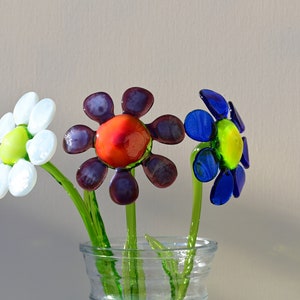 May include: Three glass flowers in a clear glass vase. The flowers are white, purple, and blue. The white flower has a green centre. The purple flower has a red centre. The blue flower has a yellow centre.