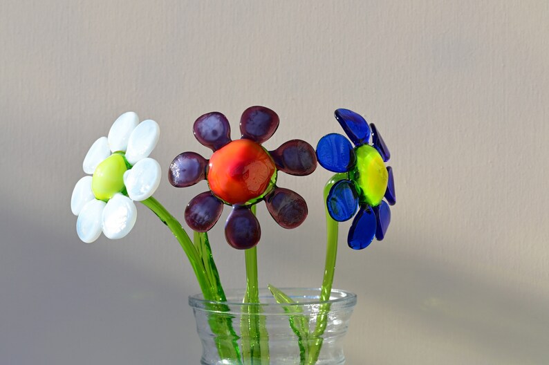 May include: Three glass flowers in a clear glass vase. The flowers are white, purple, and blue. The white flower has a green centre. The purple flower has a red centre. The blue flower has a yellow centre.