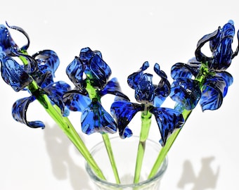 Puede incluir: Un ramo de cinco iris de vidrio azul con tallos verdes en un jarrón de vidrio transparente. Los iris están hechos de vidrio soplado y tienen un diseño delicado e intrincado.