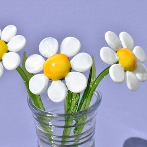 May include: Three white glass daisies with yellow centers in a clear glass vase. The vase has a textured design.