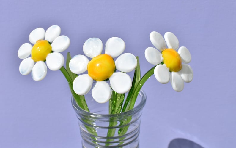 May include: Three white glass daisies with yellow centers in a clear glass vase. The vase has a textured design.