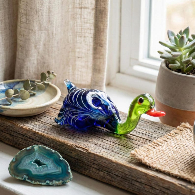 May include: A glass duck figurine featuring a deep blue body with swirling white patterns, a bright green neck, and a small orange beak. The duck is positioned on a rustic wooden surface alongside a teal agate slice and a ceramic dish.