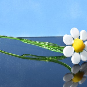 May include: A glass daisy flower with a white center and yellow center, with a green stem. The flower is on a reflective surface.