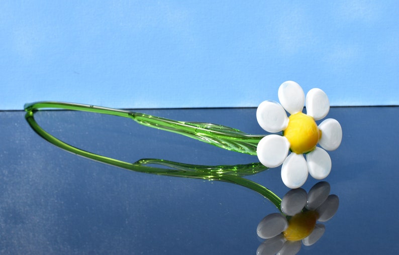 May include: A glass daisy flower with a white center and yellow center, with a green stem. The flower is on a reflective surface.