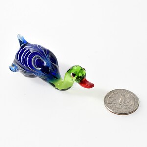 May include: A small glass duck figurine featuring a vibrant blue body with white swirling patterns, a light green neck and head, and a bright red beak. The duck is positioned next to a standard United States quarter coin for scale reference.