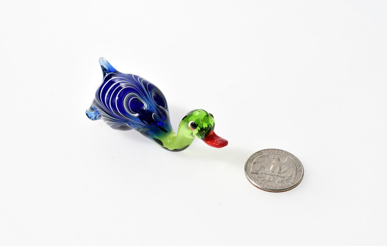 May include: A small glass duck figurine featuring a vibrant blue body with white swirling patterns, a light green neck and head, and a bright red beak. The duck is positioned next to a standard United States quarter coin for scale reference.