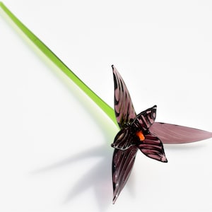 May include: A handcrafted glass flower with a dark purple bloom and a bright green stem. The flower's petals have a striped pattern, and the center features an orange accent. The stem is long and slender, and the flower is set against a white background.