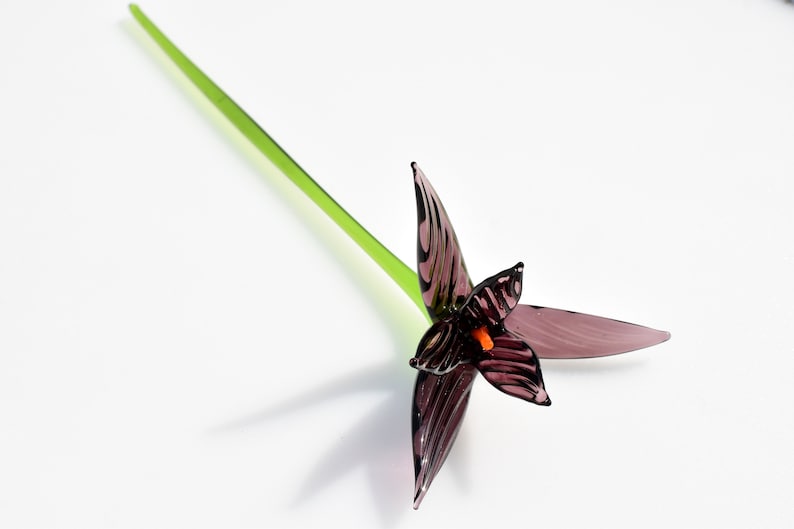 May include: A handcrafted glass flower with a dark purple bloom and a bright green stem. The flower's petals have a striped pattern, and the center features an orange accent. The stem is long and slender, and the flower is set against a white background.