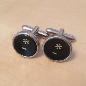 May include: A pair of silver cufflinks with black round face and a white asterisk and a horizontal line design.