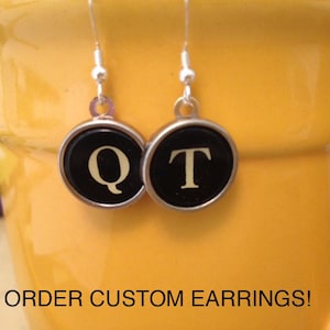 May include: A pair of earrings with black and silver accents. Each earring features a round, black button with a gold letter, one with the letter "Q" and the other with the letter "T". The earrings are hanging from a yellow ceramic mug. The text "ORDER CUSTOM EARRINGS!" is printed on the mug.