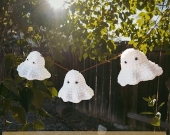 May include: A string of three white crochet ghosts with black eyes hanging from a brown string. The ghosts are suspended in front of a green leafy background with the text "PDF CROCHET PATTERN" at the bottom.