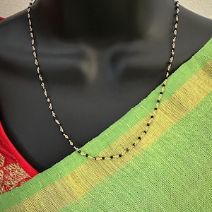 Puede incluir: Un collar de cadena dorada con cuentas negras. El collar está en un maniquí negro que lleva un sari verde y dorado.