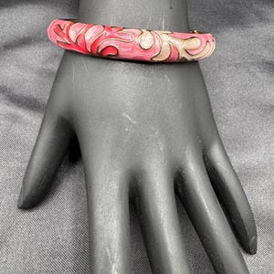 May include: A pink and red enamel bangle bracelet with a floral design. The bracelet is on a black hand. The bracelet has gold accents and a clasp.