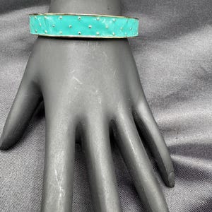 May include: A turquoise enamel bangle bracelet with a gold-tone trim and small, evenly spaced silver-tone dots. The bracelet is displayed on a black hand against a dark gray background. This piece of jewelry is a stylish accessory.