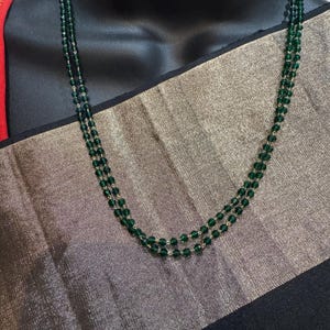 May include: A two-strand necklace featuring small, round, dark green beads. The necklace is displayed on a black mannequin, with a gold and black patterned fabric in the background.