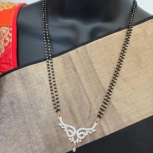 May include: A black beaded necklace with a gold-colored pendant. The pendant is shaped like a stylized wing design and is encrusted with clear gemstones. The necklace is displayed on a black mannequin.