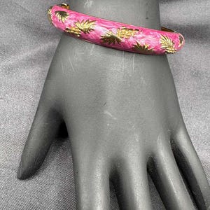 May include: A pink bangle bracelet with gold-colored palm tree designs. The bracelet is on a black hand. The bracelet has a gold-colored clasp.