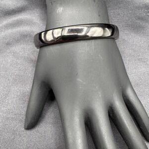 May include: A shiny, silver-colored bangle bracelet. The bracelet is a solid band with a smooth, reflective surface. The bracelet is displayed on a mannequin hand.