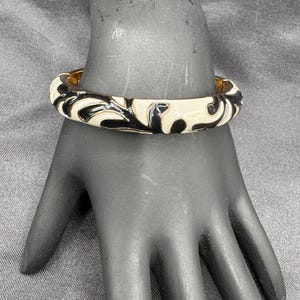 May include: A black and white enamel bangle bracelet with a gold-tone metal band. The bracelet features a repeating pattern of black swirls on a cream-colored background. The bracelet is displayed on a black hand.