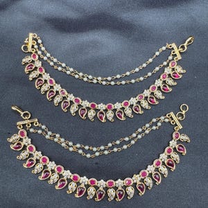 May include: Two sets of gold-coloured ear chains featuring ruby-red teardrop-shaped stones and small white gems. Each chain has a strand of small white beads. The jewellery is displayed on a dark grey surface.
