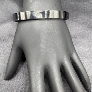 May include: A silver-colored, flat-top bracelet. The bracelet is a solid band with a smooth, reflective surface. The bracelet is shown on a mannequin hand.