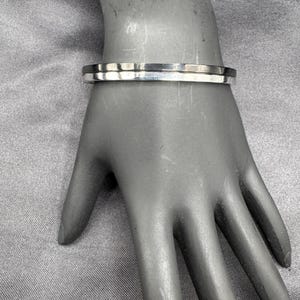 May include: A silver-colored bracelet with a flat, rectangular design. The bracelet is shown on a mannequin hand, showcasing its simple, modern style. The bracelet appears to be made of metal and has a polished finish.