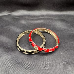 May include: Two decorative bangles. One bangle is red with white floral accents, the other is black with a gold-colored base. Both are circular and appear to be made of metal.