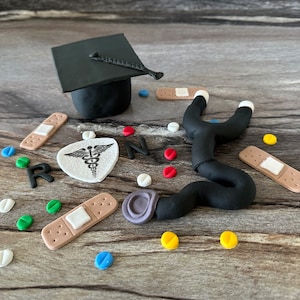 May include: A collection of handmade clay decorations, including a black graduation cap, a stethoscope, bandages, pills, and the letters "RN" and a medical symbol. The items are arranged on a wooden surface, with a variety of colors.