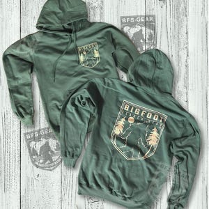 May include: A green hooded sweatshirt with a gold graphic of a Bigfoot standing in a forest with the text "Bigfoot Search Gear" on the back. The front of the sweatshirt has a smaller gold graphic of a Bigfoot in a forest with the text "Bigfoot" above it.