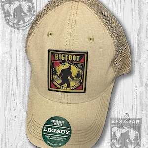 May include: A tan and brown trucker hat with a mesh back. The front of the hat features a patch with a black Bigfoot silhouette in a forest scene. The patch has the text "Bigfoot Search Gear" and "Est. 2018". The hat also has a green circle patch with the text "Dashboard Trucker Legacy".