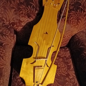 Puede incluir: Una lira de arco de madera clara, con clavijero en forma de corazón y un arco apoyado sobre las cuerdas. El instrumento tiene una forma única con un cuerpo central y lados curvos. El fondo es una tela estampada.