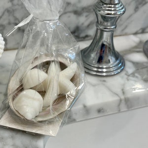 May include: A clear cellophane bag contains a seashell and white soap shaped like seashells and a starfish. The bag is tied with a white ribbon. A chrome faucet is in the background.