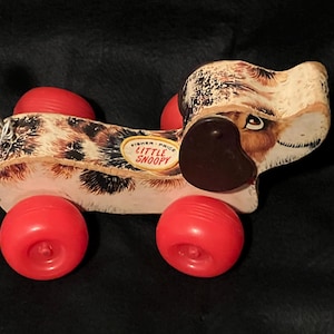 May include: Vintage Fisher-Price Little Snoopy pull-along toy dog. The toy features a spotted body, brown ear, and red wheels. The toy has a green pull string and a Fisher-Price label.