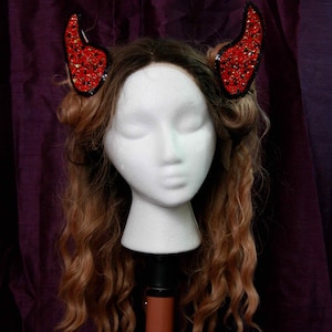 Könnte beinhalten: Ein rotes und schwarzes Teufelshorn-Haarband mit Strasssteinen. Das Haarband befindet sich auf einem Mannequin mit langen, welligen braunen Haaren.