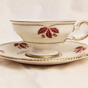 May include: Cream-colored teacup and saucer set with gold trim and a burgundy leaf design. The teacup has a handle and sits on a pedestal base. The saucer has a matching leaf pattern and gold edging. A classic tea set.