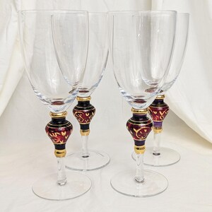 May include: Four clear wine glasses with ornate stems. The stems feature a gold band, a black section, and a burgundy bulbous design with gold accents. The glasses are displayed on a white surface.