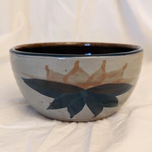 May include: A ceramic bowl with a dark brown rim and a light blue-grey exterior. The bowl is decorated with a stylized floral design in shades of brown and dark teal. The bowl is sitting on a white surface.