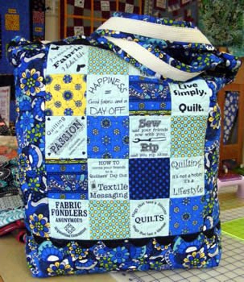 May include: A quilted tote bag featuring a patchwork design of blue, yellow, and white fabric squares. The squares display various floral patterns, polka dots, and text including "Happiness = Good fabric and a DAY OFF" and "Quilting is my PASSION."