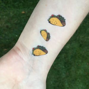 Op de afbeelding: Drie kleine, kleurrijke tijdelijke tatoeages van taco's worden op een pols getoond. De taco's hebben gele schelpen, bruine randen, groene sla, rode tomaten en bruin vlees. De tatoeages staan op een lichte huidskleur.