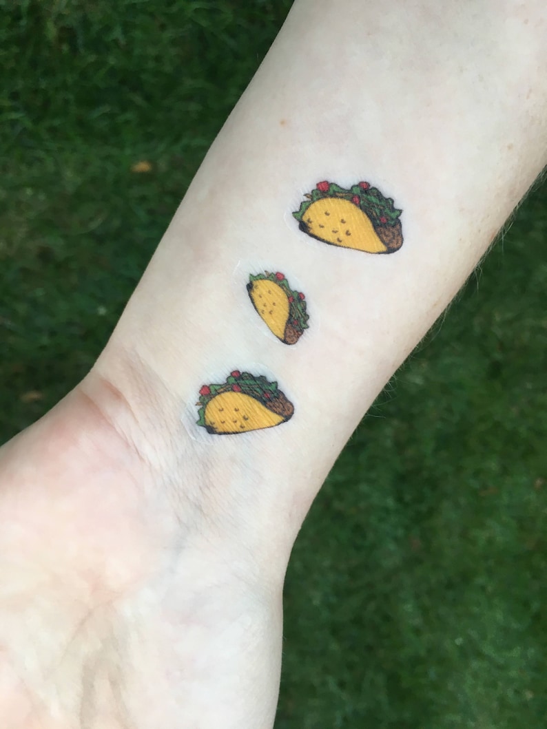 Op de afbeelding: Drie kleine, kleurrijke tijdelijke tatoeages van taco's worden op een pols getoond. De taco's hebben gele schelpen, bruine randen, groene sla, rode tomaten en bruin vlees. De tatoeages staan op een lichte huidskleur.